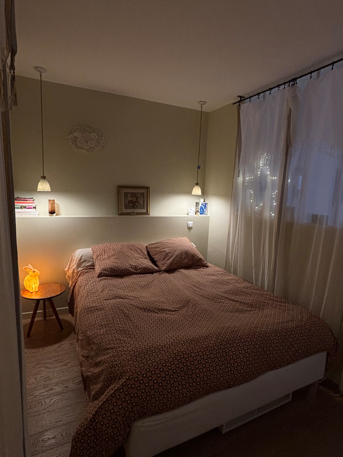 A small bedroom features a spacious bed dressed in patterned bedding. Soft light is emitted from two pendant lamps above the bed, complemented by a bedside table with an illuminated decoration. Sheer curtains allow gentle illumination from outside.