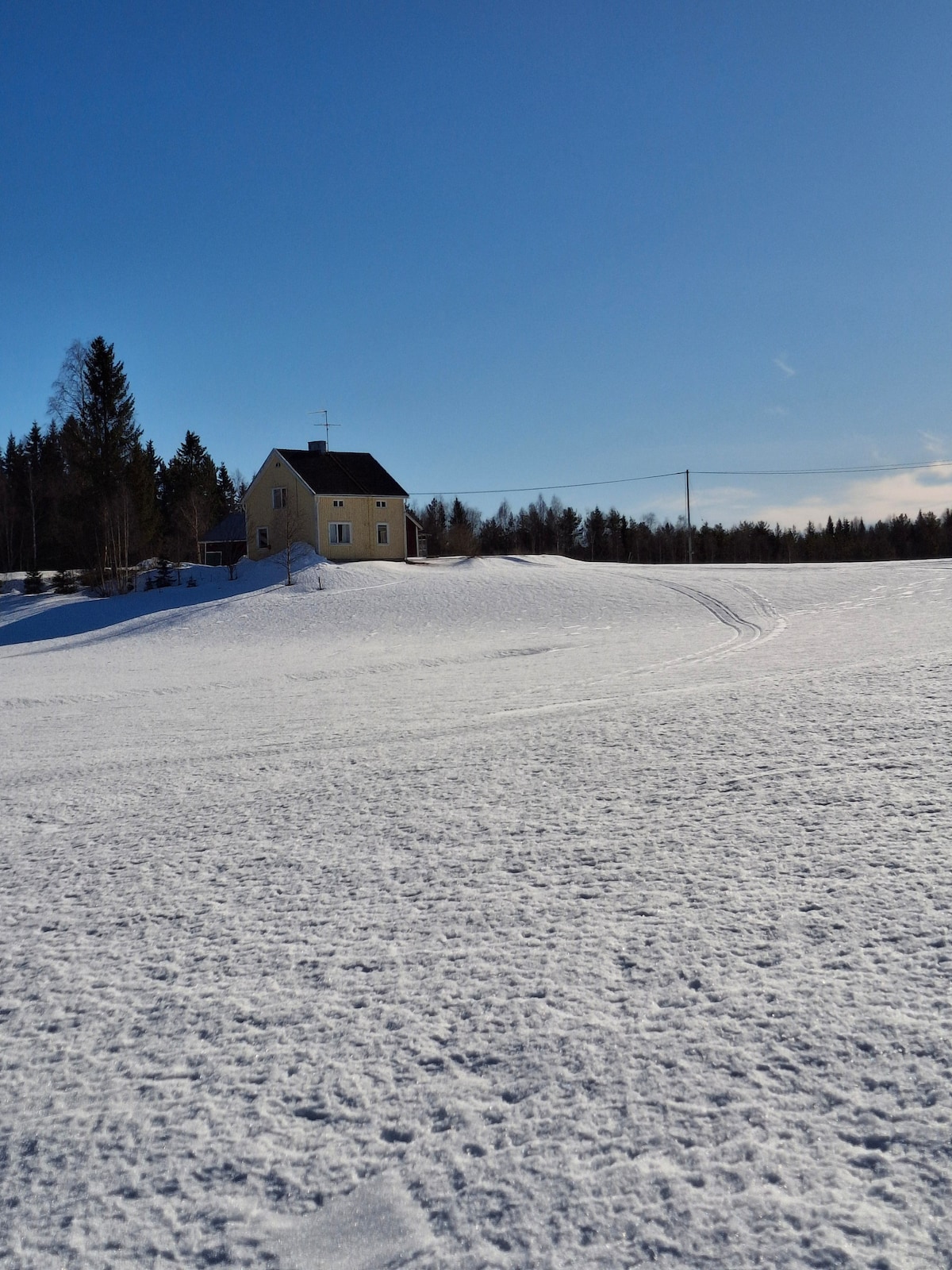 A yellow house is positioned on a snowy hillside, surrounded by a winter landscape. The clear blue sky contrasts with the white snow covering the ground, creating a serene atmosphere. Footprints and ski tracks can be seen leading towards the house.