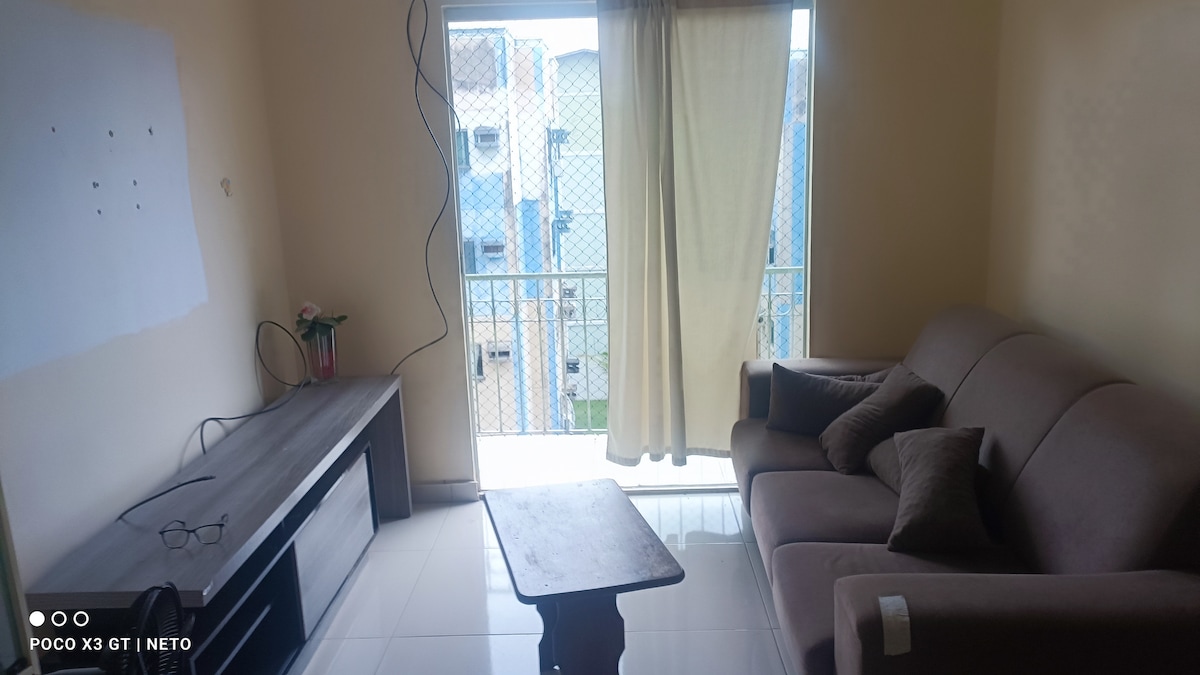 A small living area features a light-colored sofa with decorative cushions and a wooden coffee table. A television stand is positioned adjacent to a window, which has a light curtain allowing natural light to enter. A view of the outside is partially visible through the window.