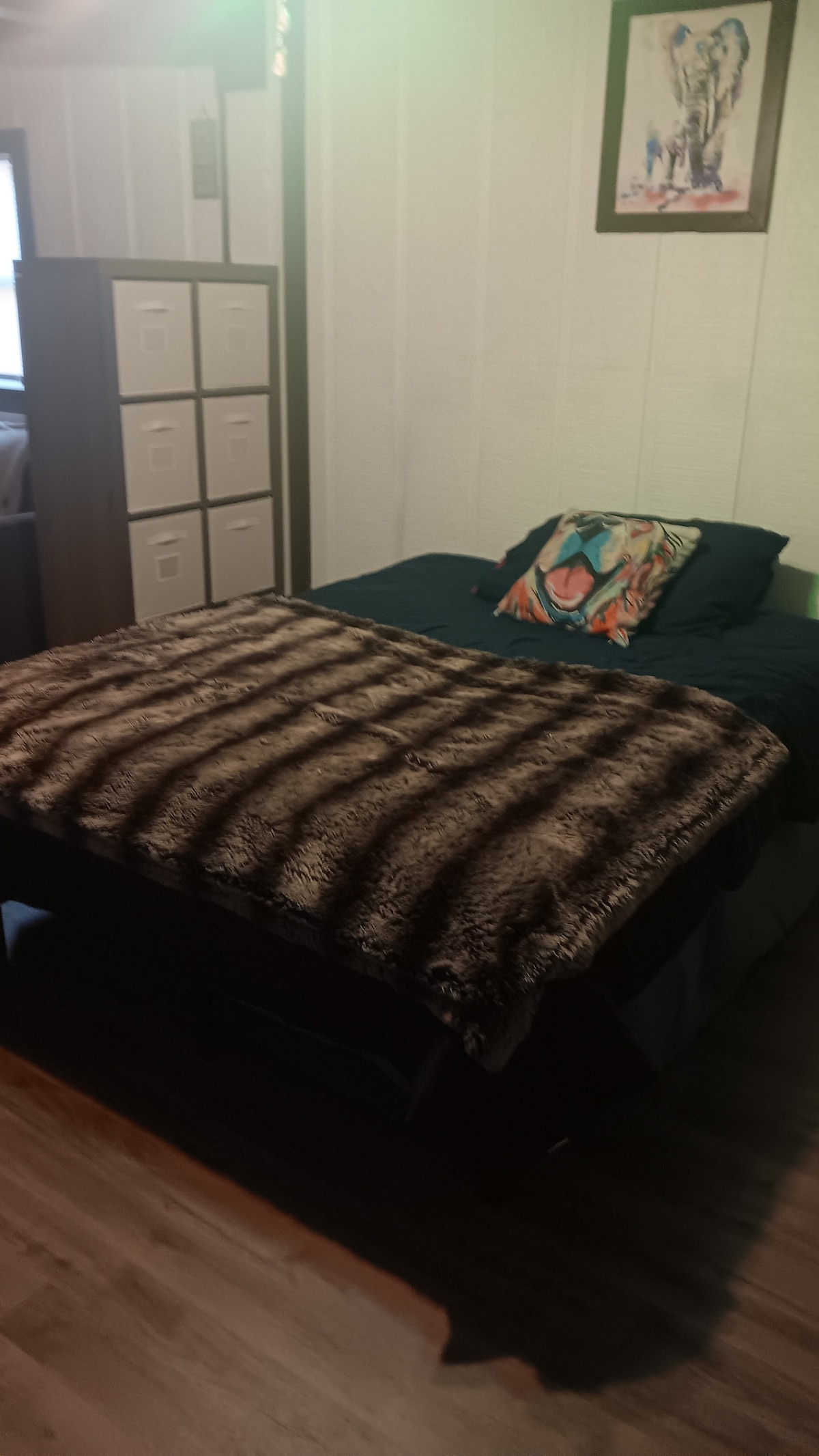 A spacious bed is draped with a soft, patterned blanket. Above the headboard, a framed piece of wall art adds a touch of color to the room. Storage units can be seen in the background, contributing to the functional layout.