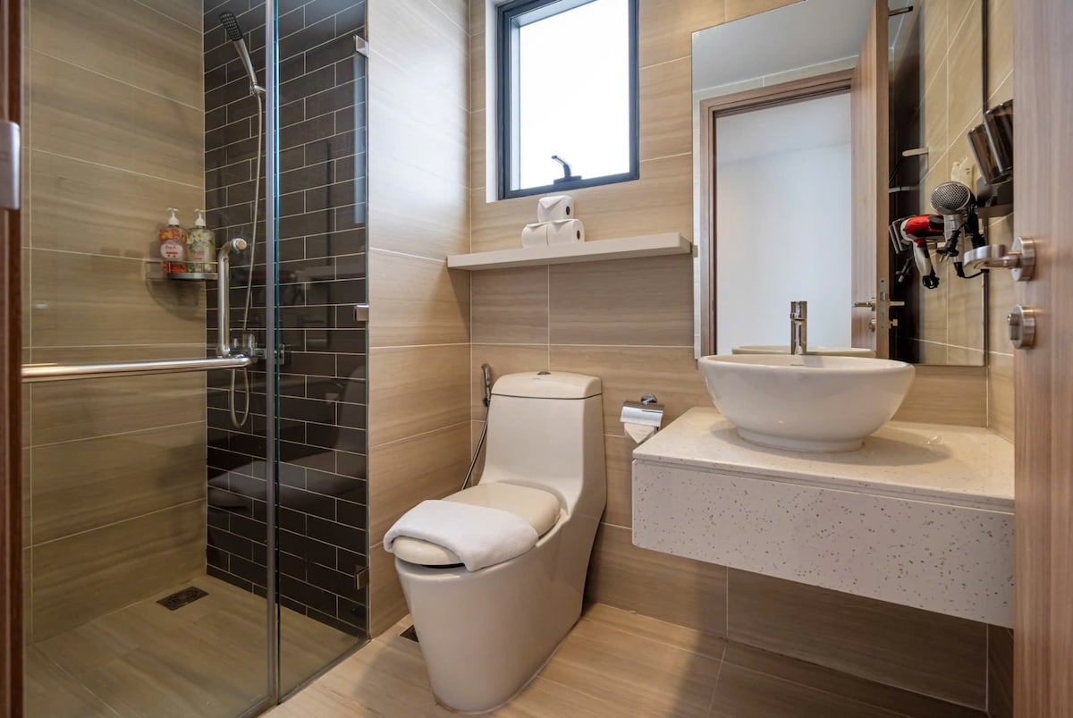 A modern bathroom is presented with sleek tile finishes in neutral tones. A walk-in shower is seen on one side, while a wall-mounted mirror is positioned above a contemporary sink. The toilet features a minimalistic design, and natural light enters through a nearby window.