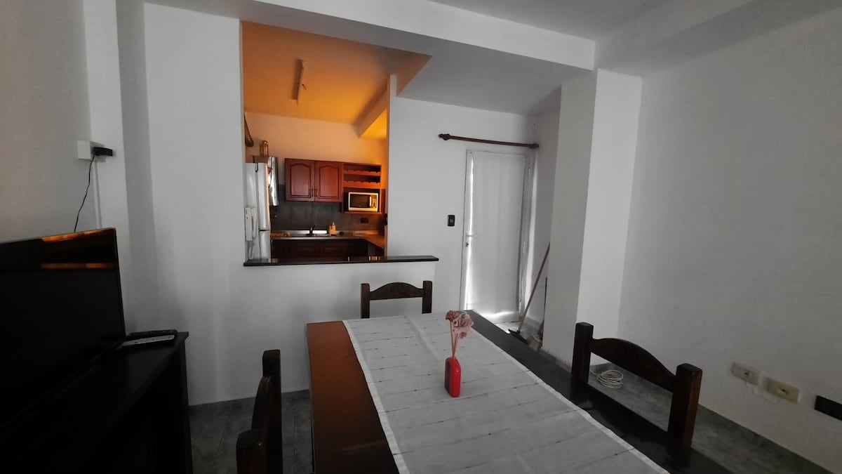 An inviting dining area is presented, featuring a rectangular table surrounded by wooden chairs. In the background, a kitchen can be seen, equipped with cabinetry and modern appliances. A small vase with a flower is centered on the table, adding a touch of color to the neutral-toned space.