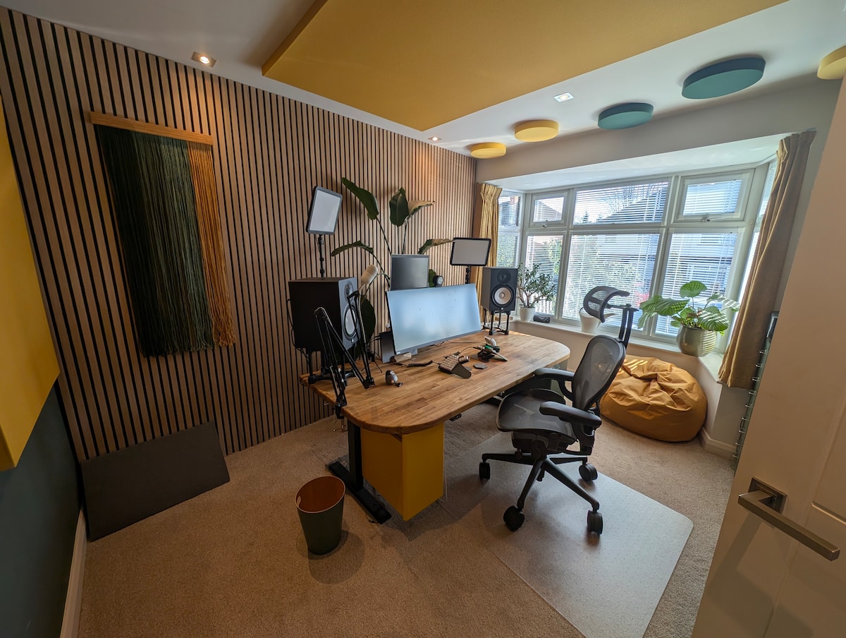 An organized workspace features a wooden desk with multiple monitors and office equipment. Soft carpet covers the floor, and natural light floods in through large windows adorned with curtains. Greenery from potted plants adds a touch of nature to the modern decor.