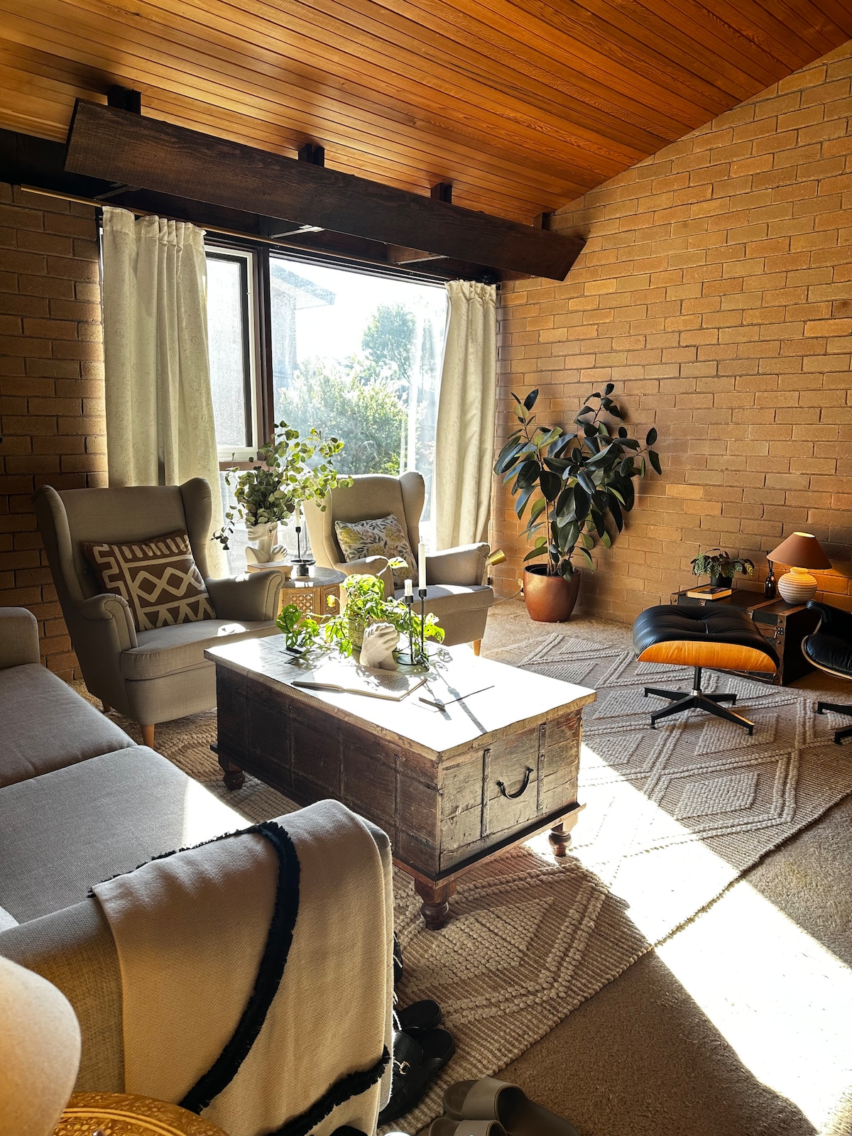 A bright sitting area is illuminated by natural light streaming through a large window. Two comfortable armchairs are positioned thoughtfully near a wooden coffee table, surrounded by potted plants. Textured rugs cover the floor, while warm wooden ceilings add to the cozy atmosphere.