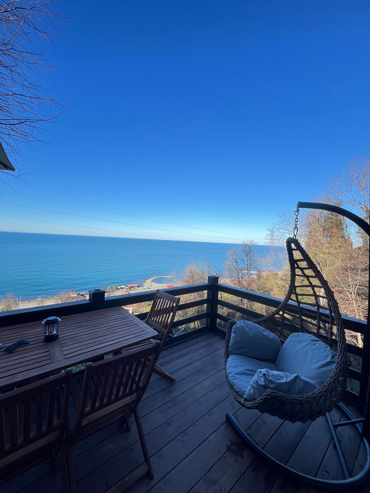 A private terrace is presented with a hanging chair and a wooden dining table surrounded by chairs. The expansive view reveals the sea, showcasing calm blue waters and a clear sky. Nearby, trees provide subtle greenery, enhancing the outdoor experience.