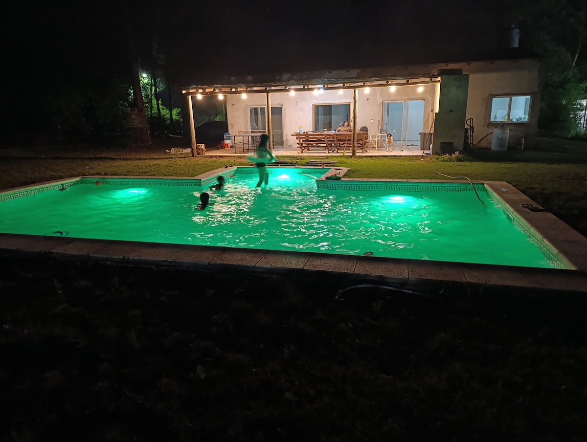 A swimming pool is surrounded by a well-maintained grassy area, illuminated with soft green lights. The house features a covered outdoor dining space in the background, with several tables and chairs visible. The atmosphere conveys a sense of relaxation and leisure.