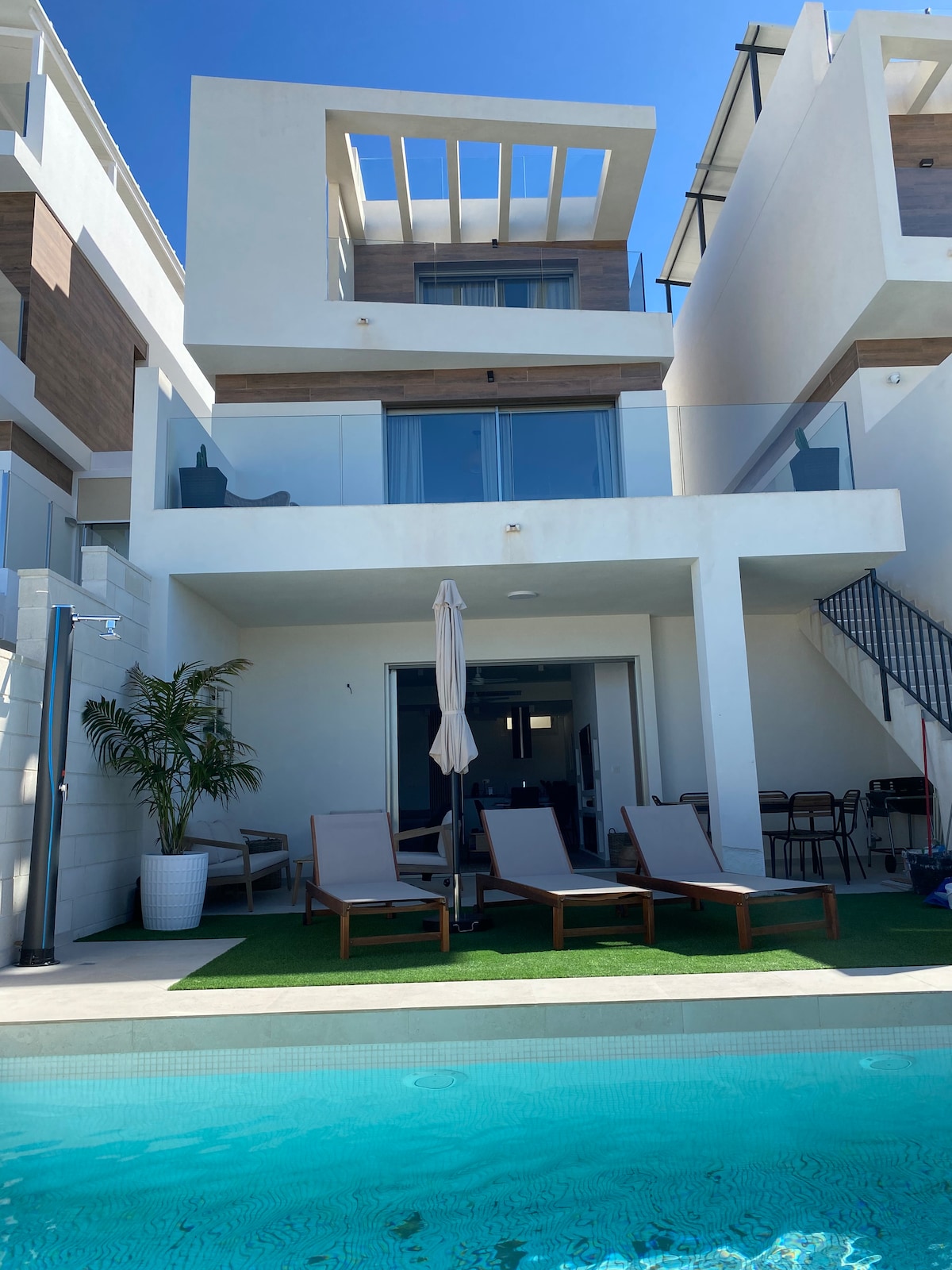 A modern villa with a clean architectural design is featured. A private pool is visible in the foreground, complemented by several lounge chairs. The upper levels have balconies, with sliding glass doors providing access to indoor spaces, surrounded by a well-maintained exterior area.