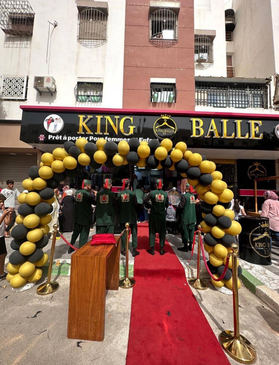 An entrance adorned with a decorative arch of black and gold balloons leads to a venue marked 'KING BALLE'. A red carpet is laid out, flanked by velvet ropes, while several individuals dressed in green stand near the entrance, welcoming guests.