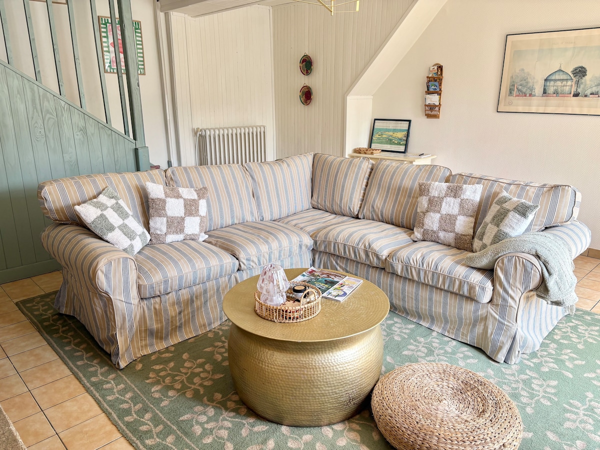 A spacious lounge area features a large striped sofa with decorative cushions, perfectly positioned around a round gold coffee table. A textured rug adds warmth to the tiled floor, while light from a window illuminates the bright, inviting space.