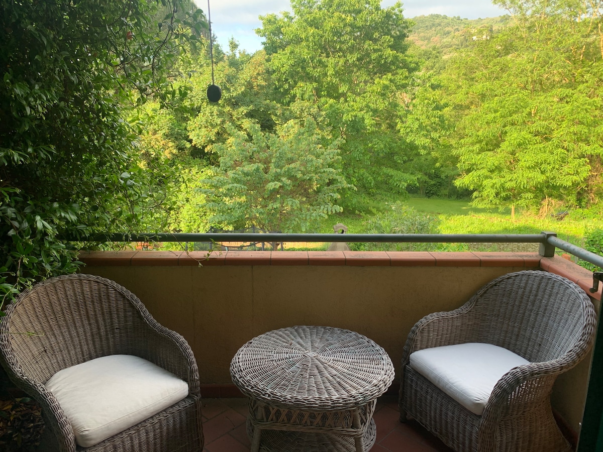 A serene outdoor veranda is furnished with two wicker chairs and a round table. Lush greenery surrounds the space, providing a peaceful view of the natural landscape. The area is accented by gentle morning light filtering through the trees.