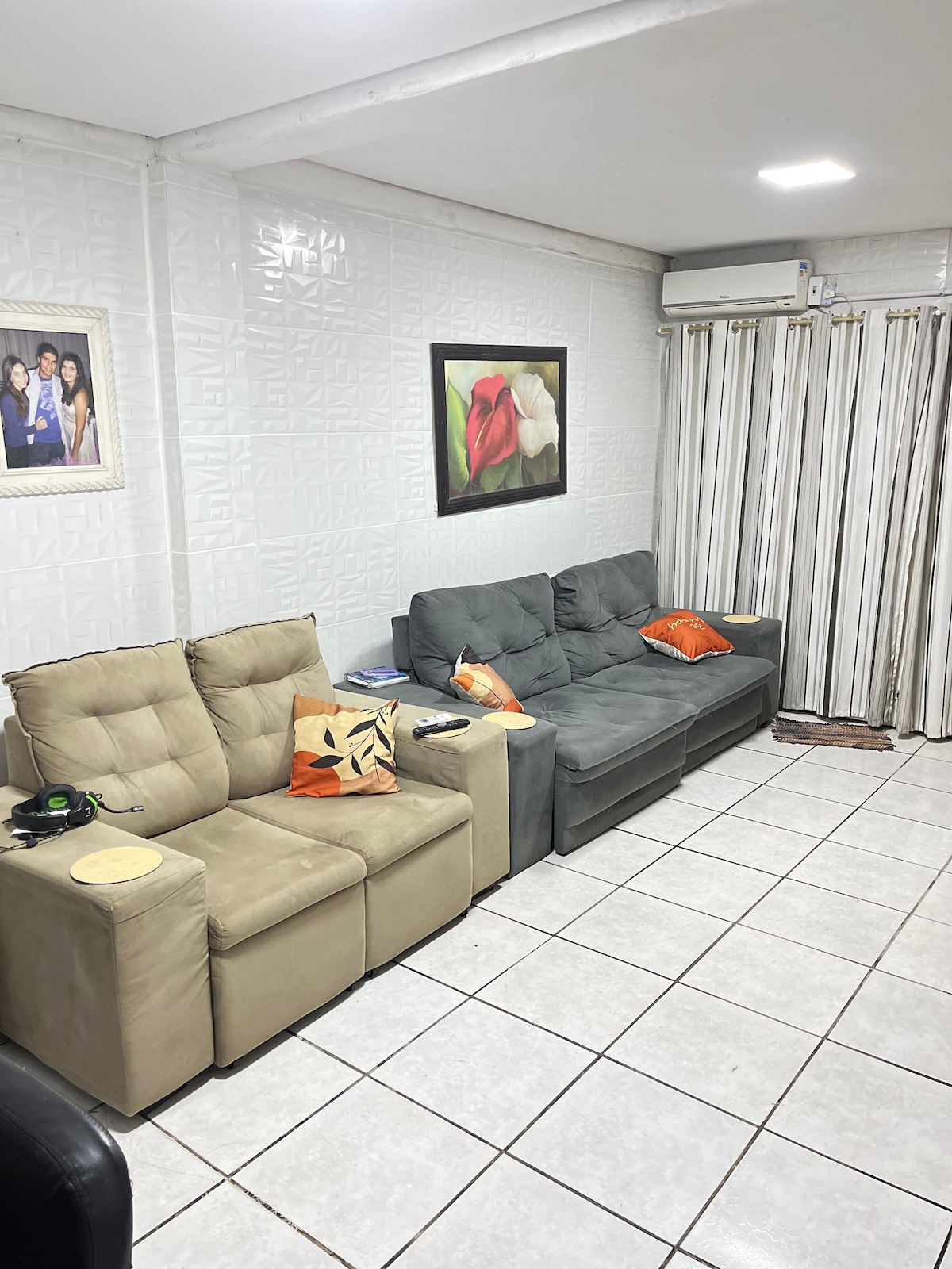 A living room is presented with three sofas of varying colors, complemented by decorative pillows. A painting featuring flowers is displayed on the wall, while light-colored tiles cover the floor. Curtains frame a window, allowing natural light to fill the space.