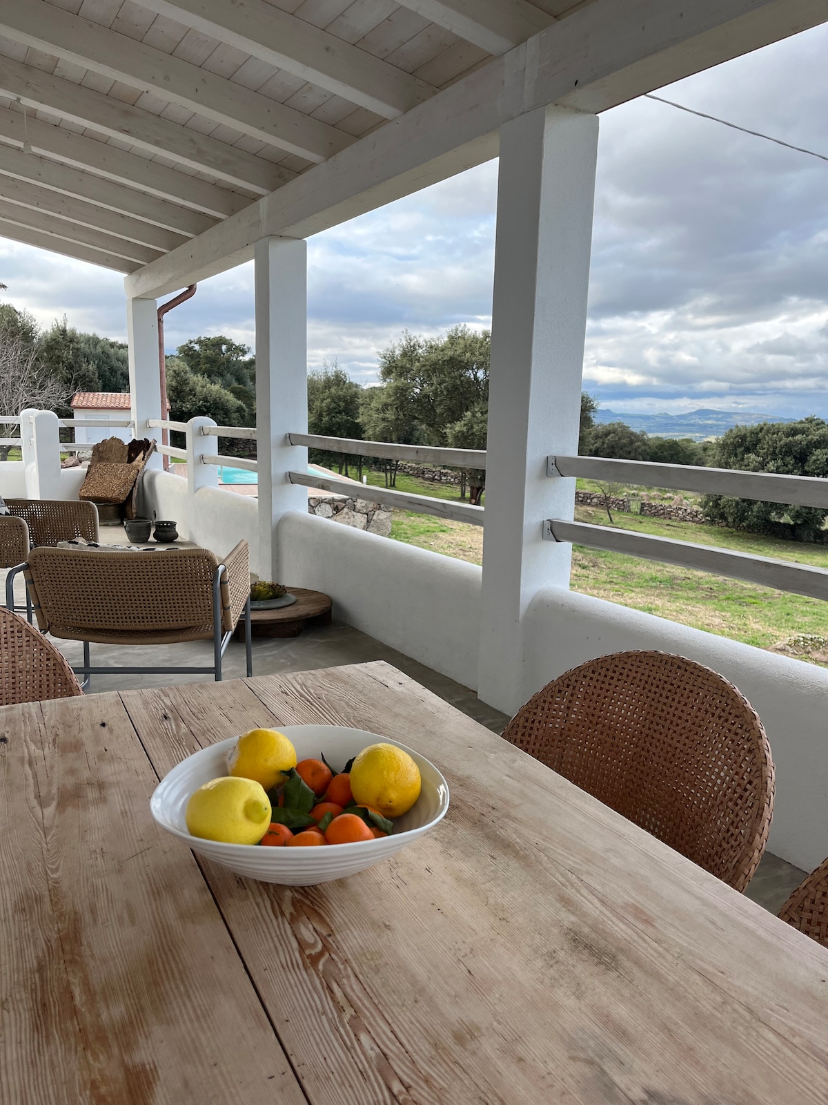 A covered veranda features a wooden dining table with a bowl of fresh fruits, including lemons and oranges. Woven chairs are positioned around the table, with distant views of the garden and surrounding countryside visible through the railing.