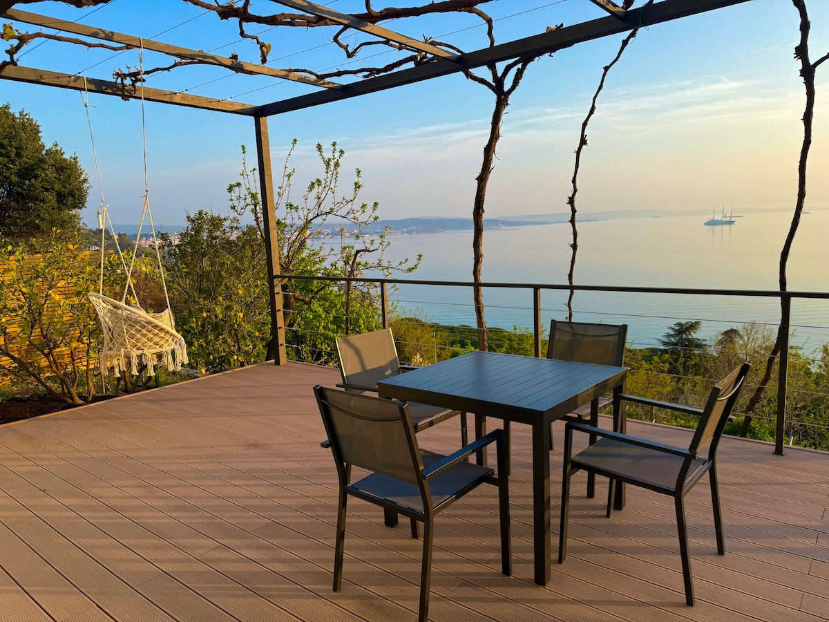 An outdoor terrace is presented with a dining table and four chairs. A swing hangs from a wooden pergola adorned with climbing plants. The view extends over the tranquil waters of the Gulf of Trieste, with gentle colors from the sunset reflecting in the distance.
