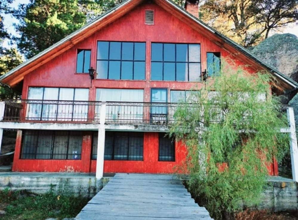 A two-story red house features large glass windows, allowing natural light to fill the interior. A wooden walkway leads to the entrance, bordered by greenery. The structure displays a combination of rustic charm and modern design, with a balcony providing outdoor views.