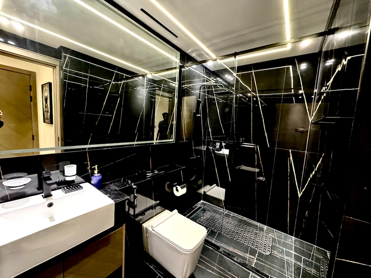 The contemporary bathroom features sleek black tiles with contrasting geometric patterns. A spacious vanity is equipped with a sink and modern fixtures, while ample mirrors create an expansive feel. Essential toiletries and fresh towels are neatly arranged for guest convenience.