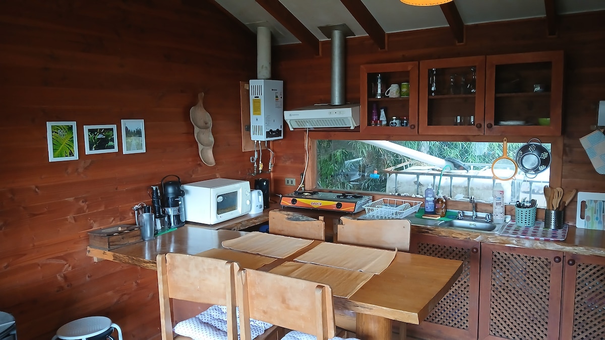 The kitchen area features wooden walls and a rustic design, equipped with essential appliances including a microwave, stove, and sink. A dining table with four chairs is positioned in the center, while shelves display dishware and cooking utensils. Natural light enters through a window that overlooks the outdoor space.