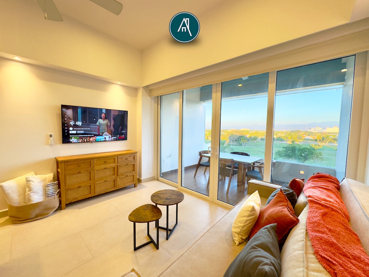 A spacious living area features large glass doors that provide a bright view of the golf course. A comfortable sofa is positioned beside a natural wood coffee table, while a television is mounted on a wooden console. Soft lighting adds to the inviting atmosphere.