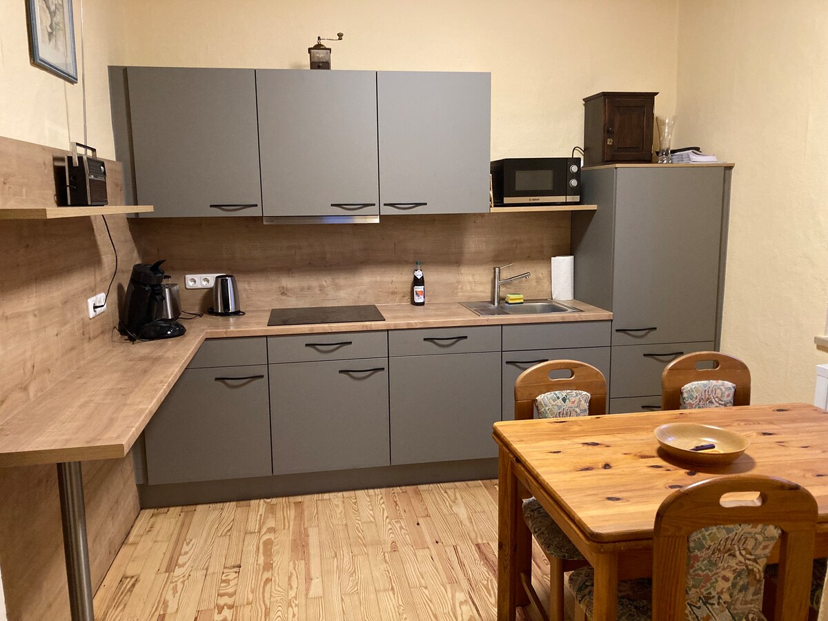 A functional kitchen area features gray cabinetry and wooden countertops. Cooking appliances, including a kettle, microwave, and stove, are visible. A wooden dining table with four chairs showcases floral upholstery. Natural wood flooring adds warmth to the space.