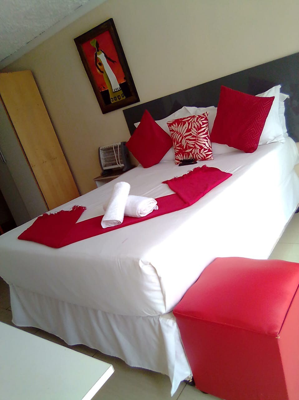 A neatly arranged bed features crisp white linens, accented by vibrant red pillows. A compact ottoman in a matching hue sits at the foot of the bed. A piece of wall art adds a touch of color, while a wardrobe is visible in the background.