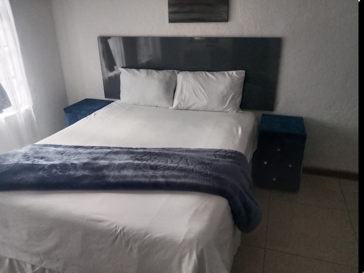 A comfortable sleeping area features a spacious bed adorned with crisp white linens. Two blue nightstands flank the bed, providing a pop of color, while a soft blue throw drapes over the end. Natural light filters in through a nearby window.