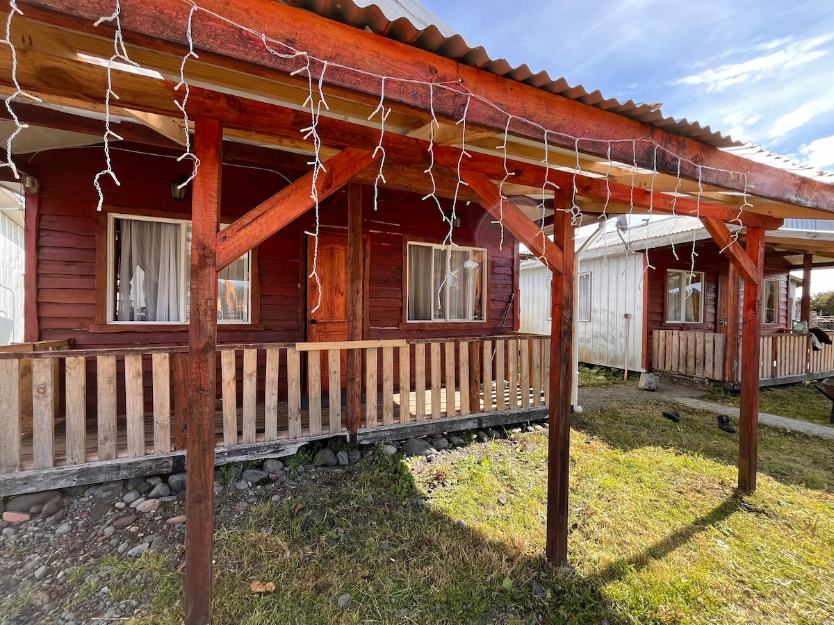 A wooden cabin is showcased, featuring a welcoming porch supported by sturdy posts. String lights are hung along the edge, adding visual interest. Two windows are visible on the front, allowing natural light to enter the interior. A grassy area surrounds the cabin.