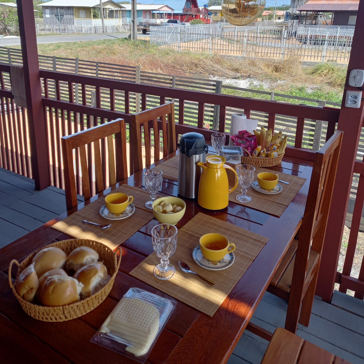 A wooden dining table is set on a porch, showcasing breakfast items. Plates of pastries, glasses of orange juice, and coffee are arranged alongside utensils. The surrounding area features wooden fences and scenic views of nearby houses.