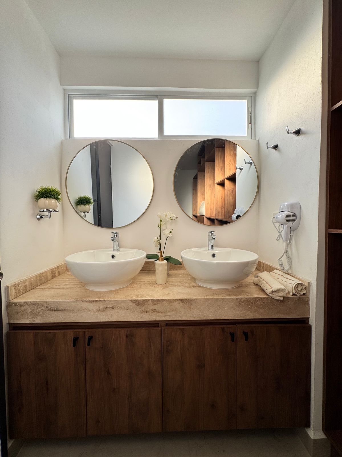 A modern bathroom features a double sink setup with round mirrors above each basin. Natural light enters through a window, highlighting the neutral-colored countertops and wooden cabinets below. A decorative plant and a small vase with an orchid add a touch of elegance.