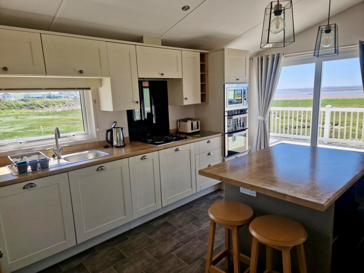 A modern kitchen features sleek cabinetry and stainless steel appliances, including a kettle and a microwave. A small wooden island with two stools provides additional seating. Large windows allow natural light to fill the space, with views of the grassy area beyond.
