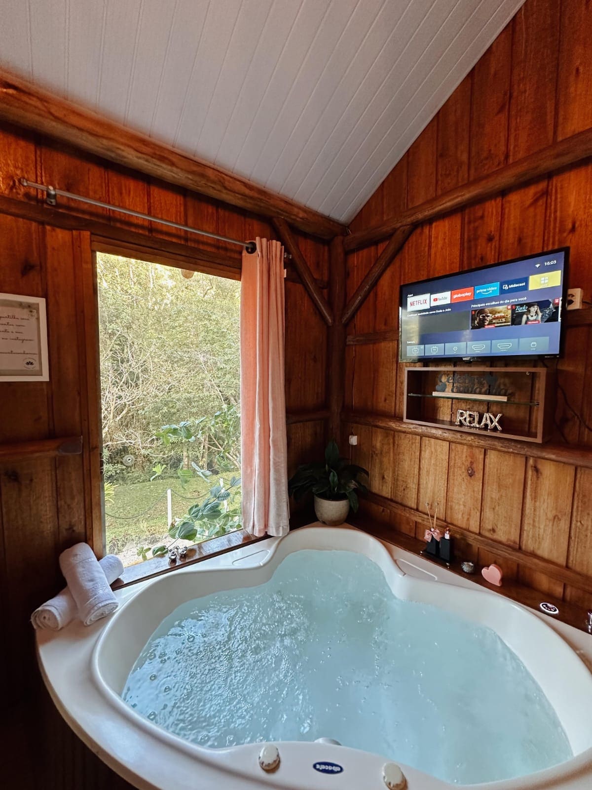 A heart-shaped hot tub is situated next to a large window, providing views of the surrounding greenery. Soft towels are neatly arranged nearby, and a wall-mounted TV is positioned above a wooden shelf with decorative items, contributing to a tranquil retreat.