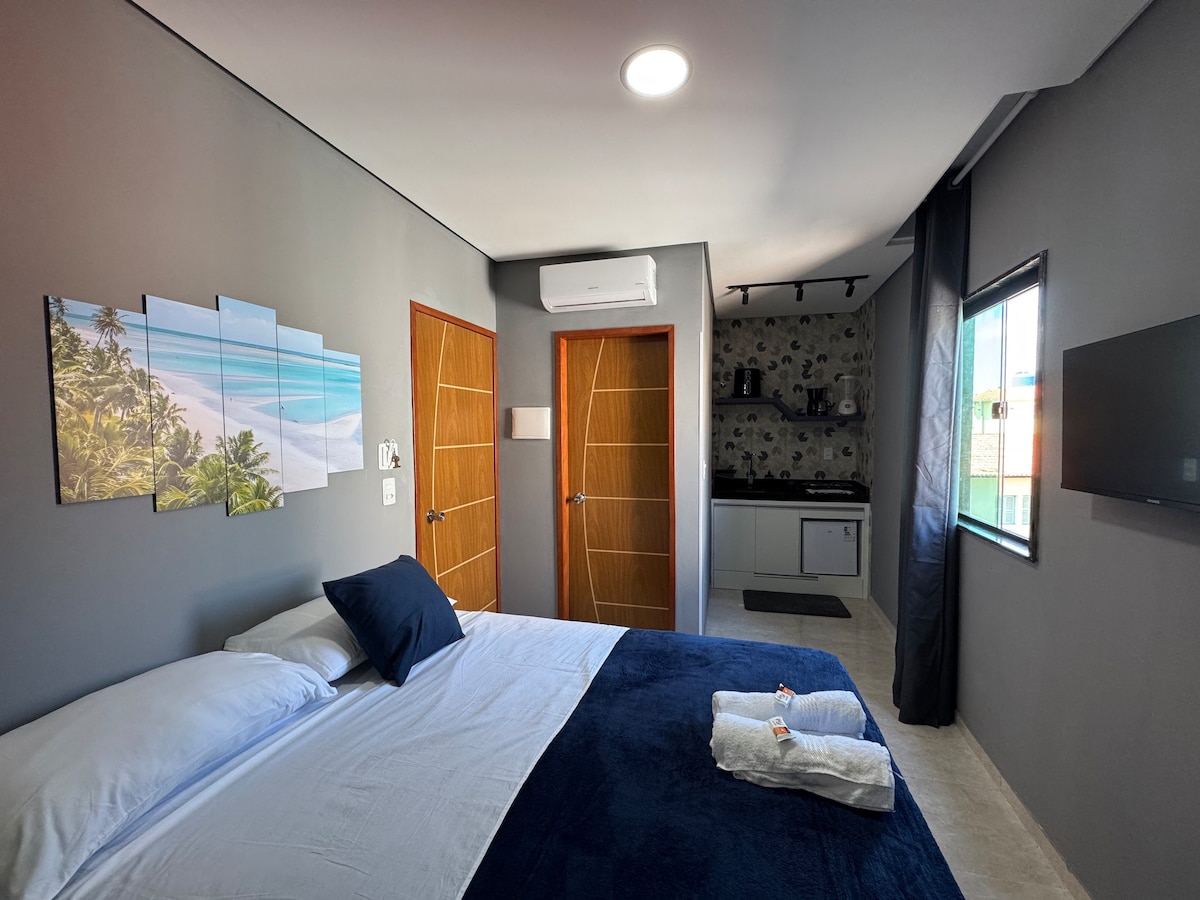 A spacious bedroom featuring a large bed with white linens and a dark blue blanket. Two doors lead to other areas, while a window with a view is covered by a dark curtain. A sleek kitchenette and a flat-screen TV are present in the corner.