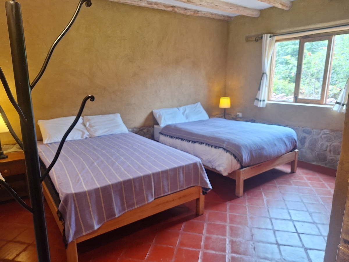 A serene sleeping area features two spacious beds, each dressed in soft linens with muted tones. Natural light enters through a window adorned with simple curtains, and warm tones of the walls are complemented by rustic wooden beams above.