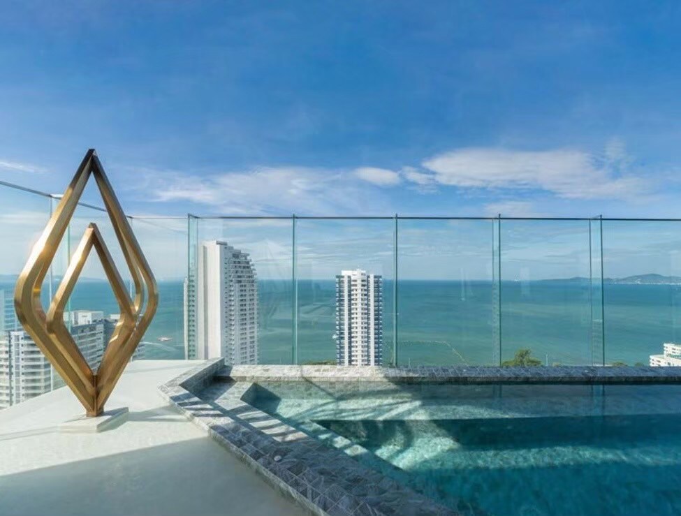 A serene outdoor pool area is showcased, featuring a glass railing that offers unobstructed views of the ocean and surrounding cityscape. A sculptural piece sits prominently, adding an artistic touch to the space while the blue sky reflects off the water's surface.