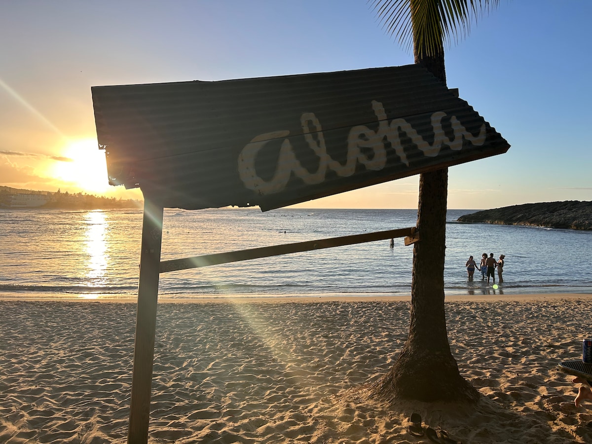 A rustic sign reading 'Aloha' is supported by a palm tree, set against a backdrop of a serene beach. The sun is setting over calm waters, casting a warm glow, while silhouettes of people can be seen wading in the water.
