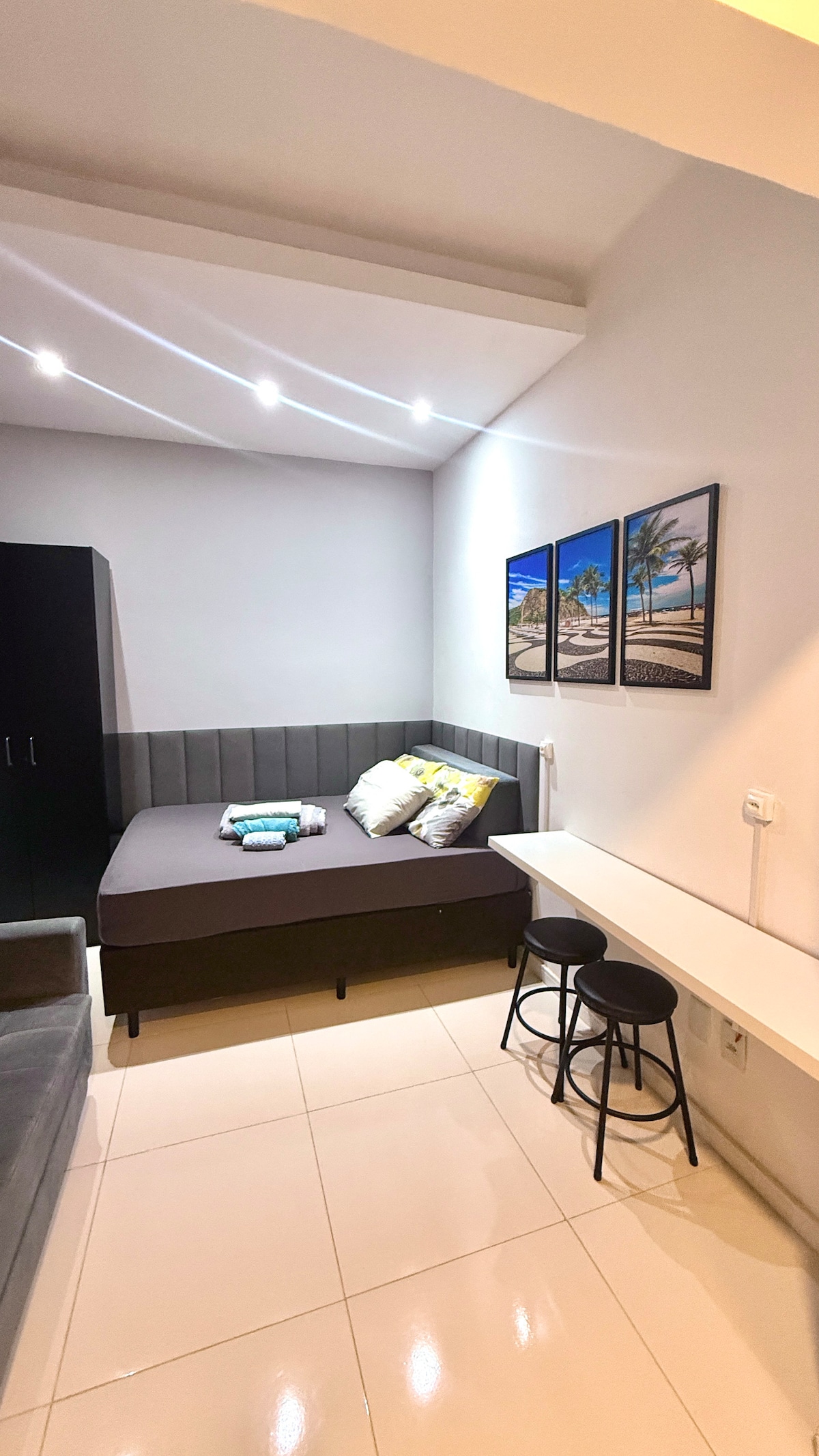 A compact living area features a spacious bed adorned with decorative cushions. Two framed beach-themed images are mounted on the light gray wall. A small table with two stools is positioned beside the bed, and a sleek black wardrobe is visible in the background.