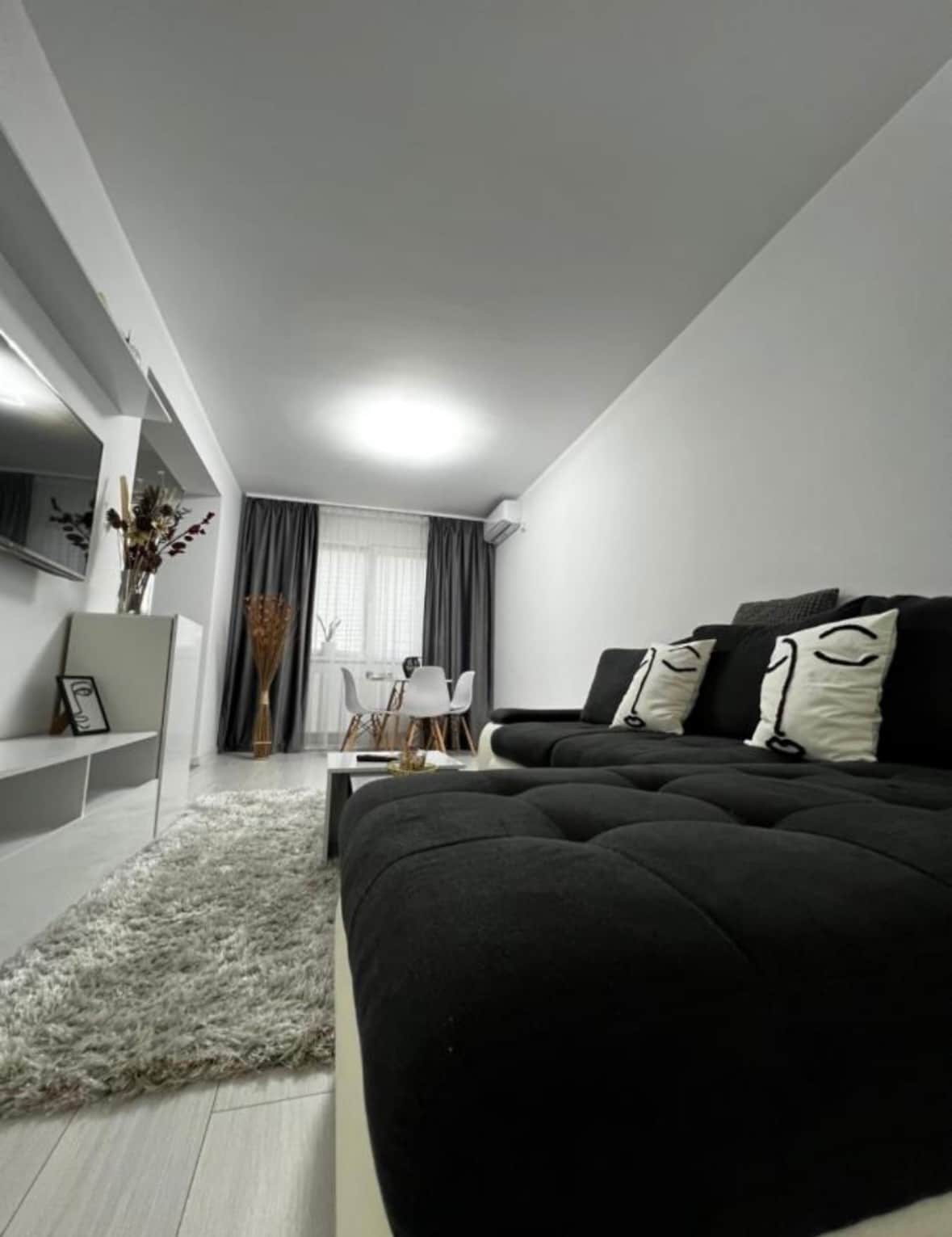 A modern living space is presented with a dark sofa adorned with decorative pillows featuring abstract faces. A soft white rug covers the floor, while a dining area with a round table and chairs is visible in the background. Natural light enters through sheer curtains.