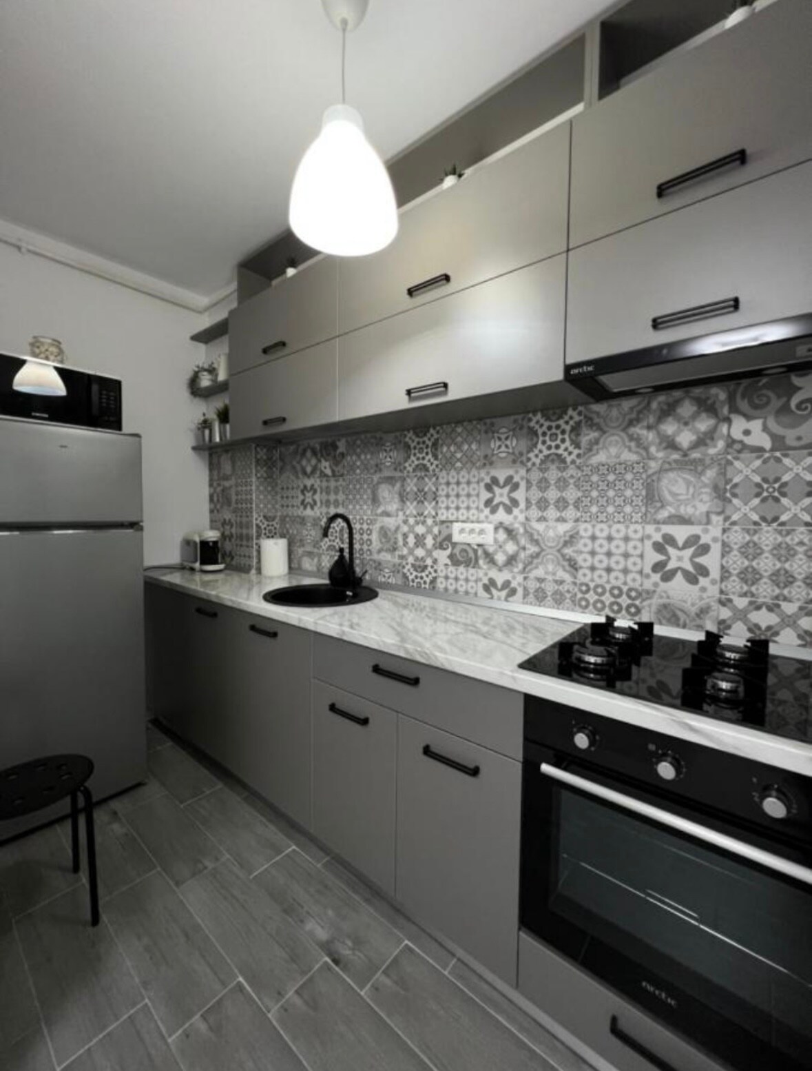 A modern kitchen is presented, featuring sleek gray cabinetry and a patterned backsplash. A refrigerator and an oven are visible, along with a stovetop. Counter space is utilized for small appliances, and a black stool is positioned near the wall.