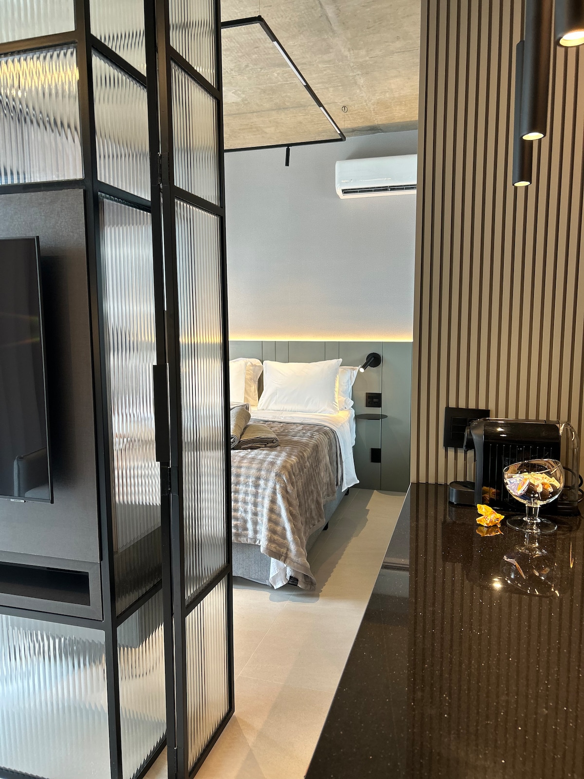 A modern bedroom is visible through a sleek glass partition, featuring a spacious bed with neutral bedding. A flat-screen TV is mounted on the wall, with ambient lighting creating a soft glow. A stylish countertop with a coffee machine is positioned in the foreground.