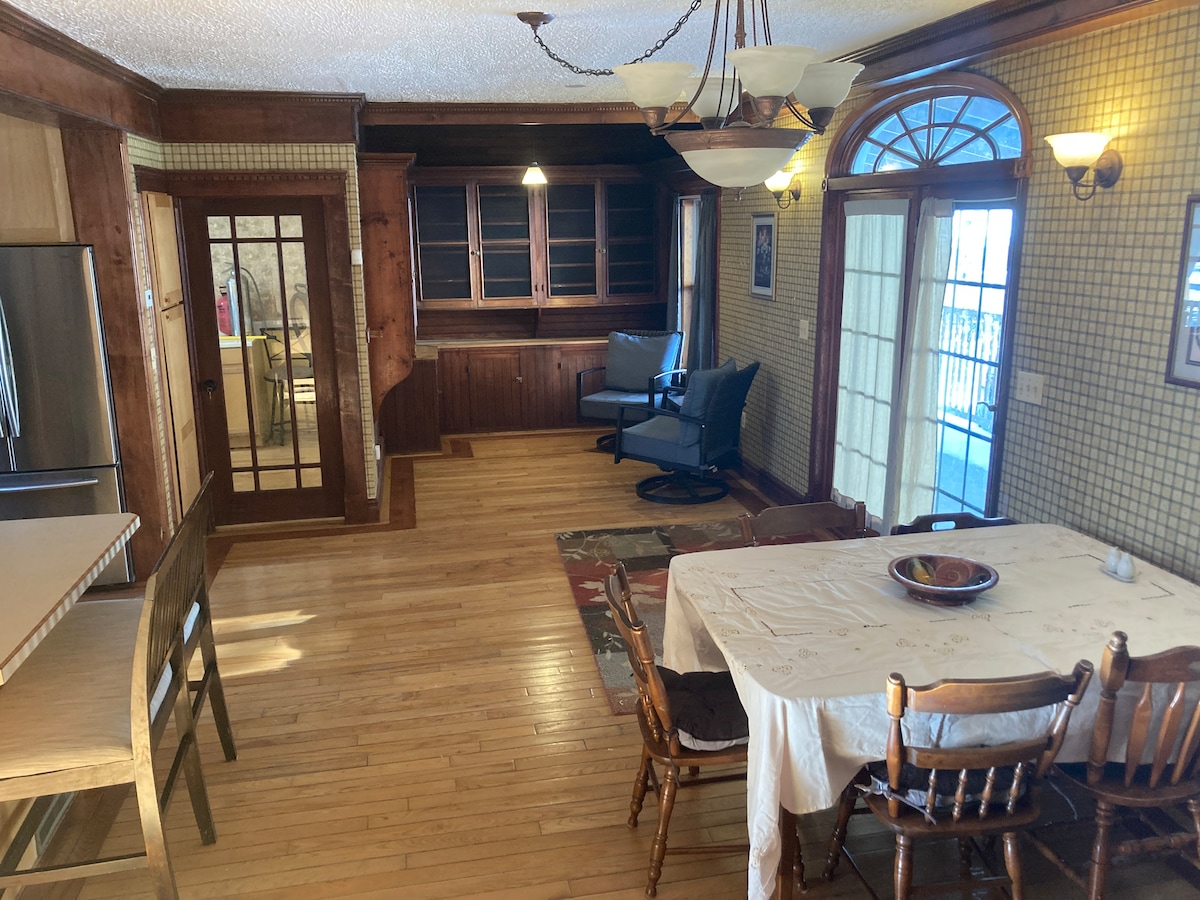 An inviting dining area is depicted with a wooden table surrounded by several chairs, set on a warm wooden floor. A buffet-style cabinet is showcased against the wall, complemented by a spacious seating area featuring a comfortable chair. Sliding glass doors open to a bright outdoor space.
