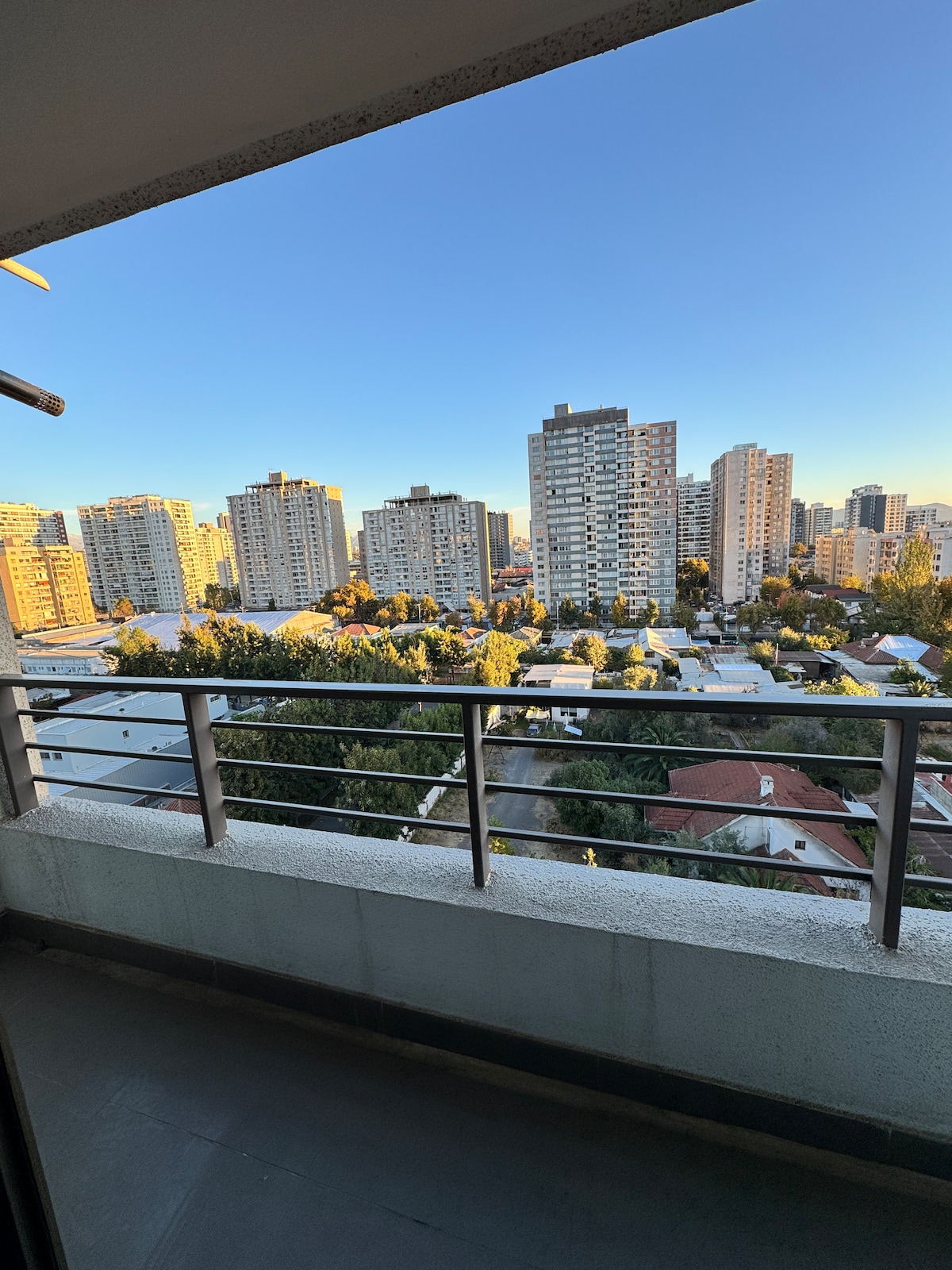 A spacious balcony offers a panoramic view of city skyscrapers and nearby residential areas, illuminated by the soft light of the early evening. Lush trees are visible among the buildings, creating a contrast against the urban landscape.