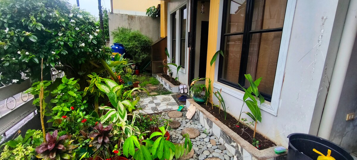 A serene pathway is lined with diverse plants and flowers, leading to a welcoming entrance. Decorative stones create a mosaic effect along the path, while the bright exterior of the building complements the greenery. A glimpse of the front door is framed by large windows.