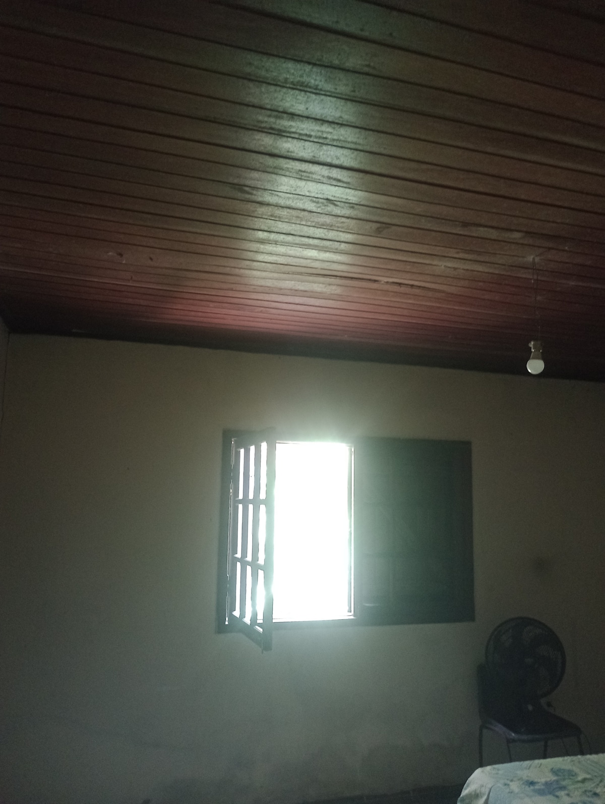 A room featuring wooden beams along the ceiling. A window, partially opened, allows natural light to enter, illuminating the space. A fan is positioned in the corner, contributing to the room's comfort.