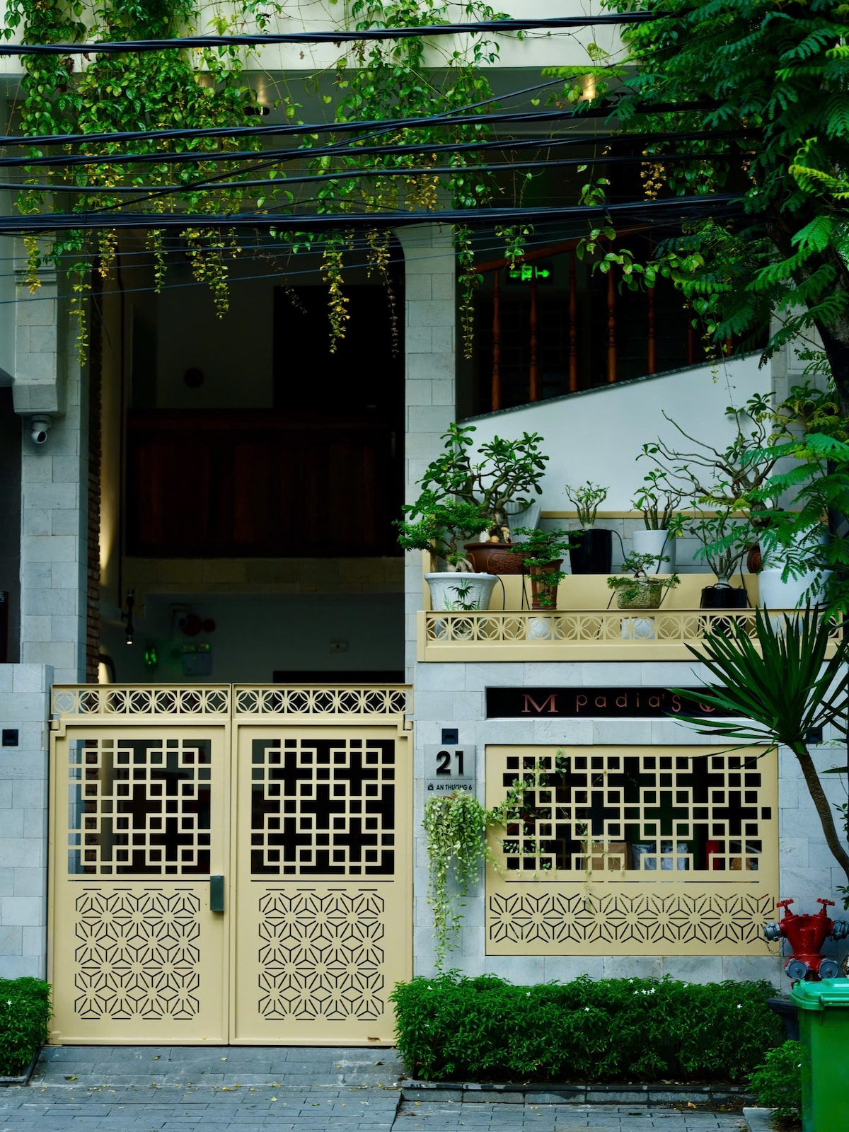 An entrance is framed by decorative golden gates, showcasing intricate designs. Lush greenery is visible, with potted plants positioned along the upper balcony. The building features a mix of stone and wooden materials, contributing to its modern style.