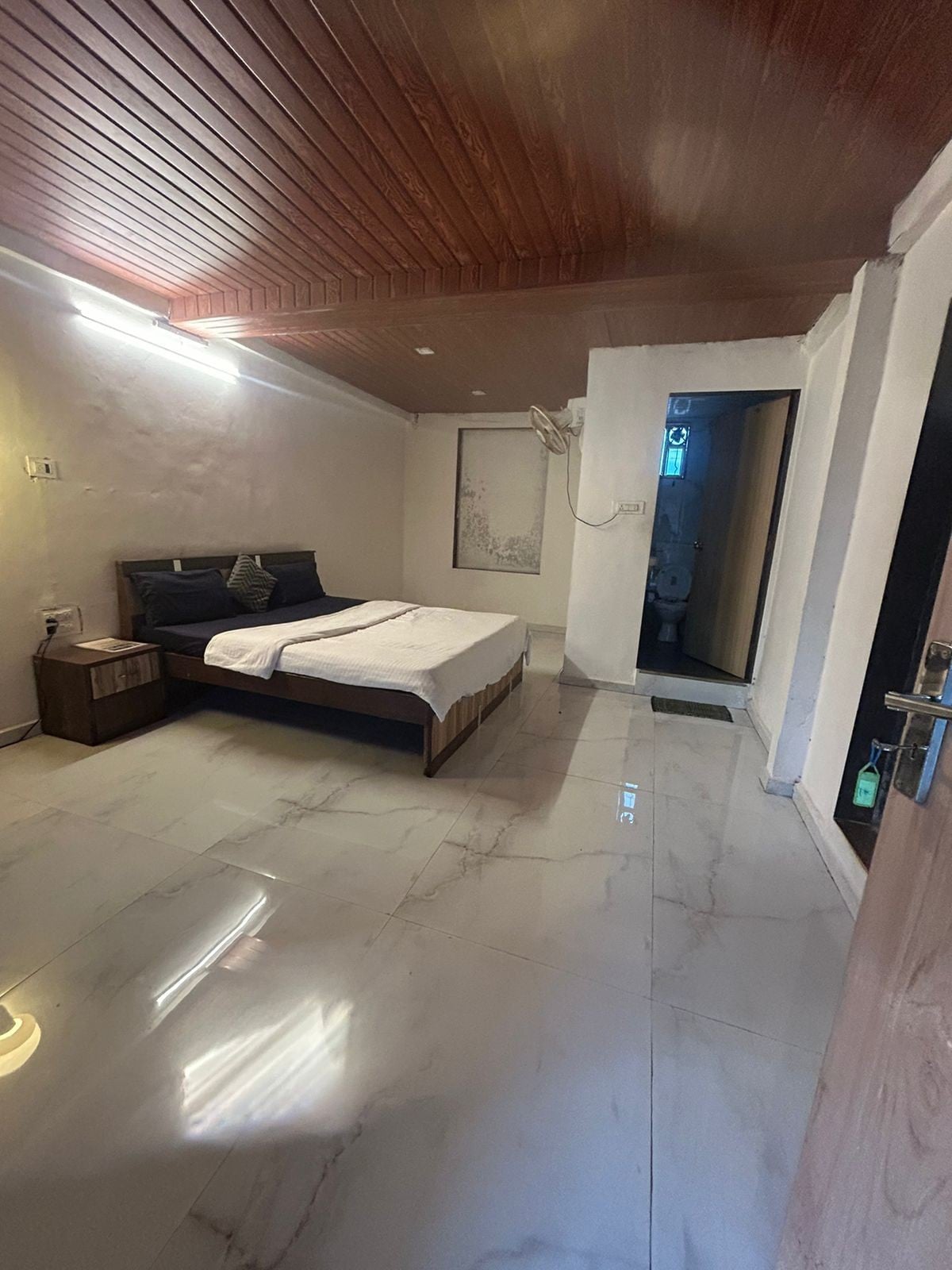 A private room features a spacious bed with dark bedding set against light-colored walls. Natural light enters through an adjacent door leading to an attached bathroom. The room is fitted with a compact nightstand and a smooth tiled floor, creating a minimalist ambiance.