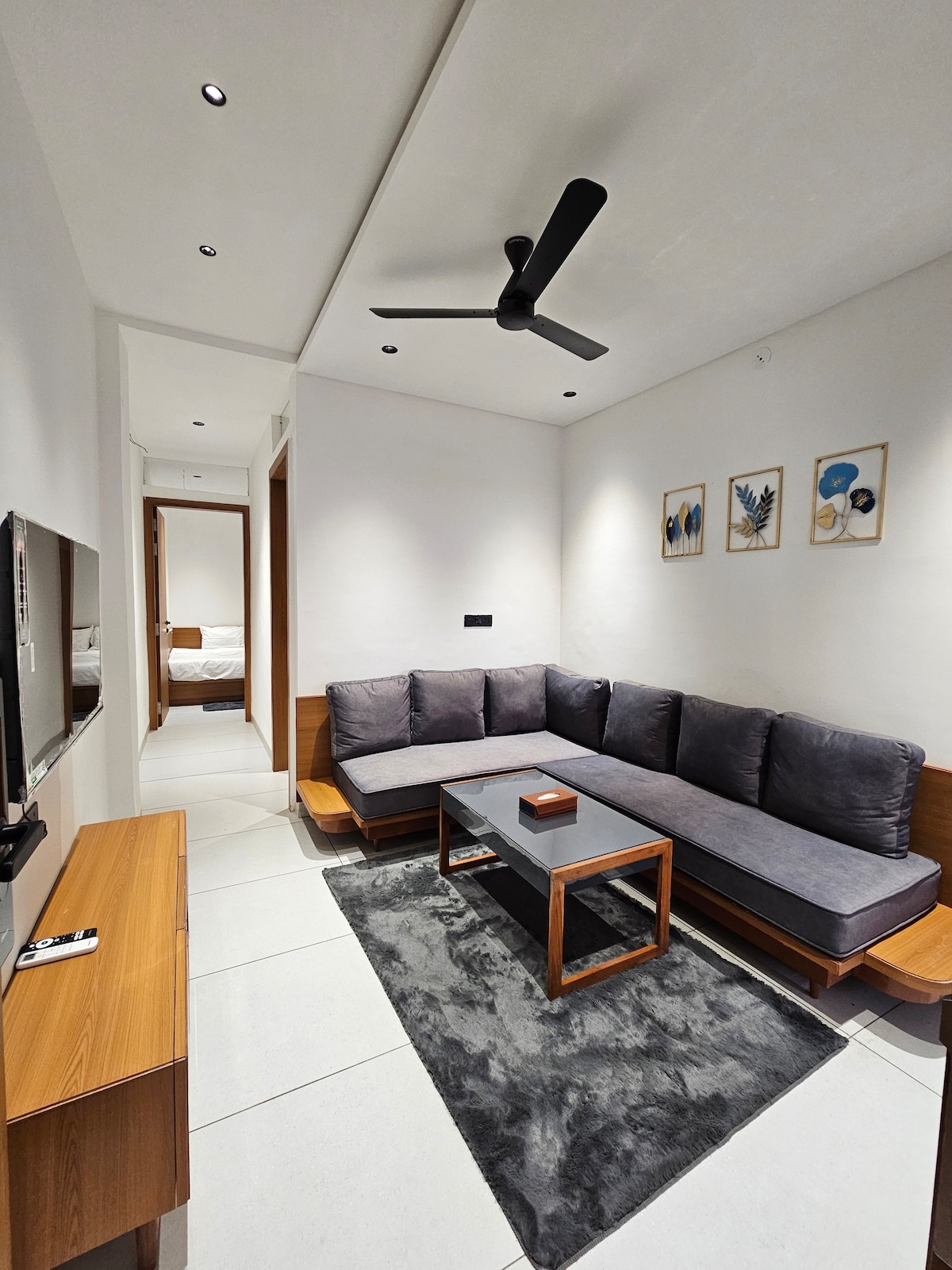 A modern living area features a cozy sectional sofa in muted gray, positioned around a sleek coffee table. Minimalistic wall art adds subtle character, and a ceiling fan provides an atmosphere of comfort. Soft light is emitted from recessed lighting, highlighting the clean lines of the space.