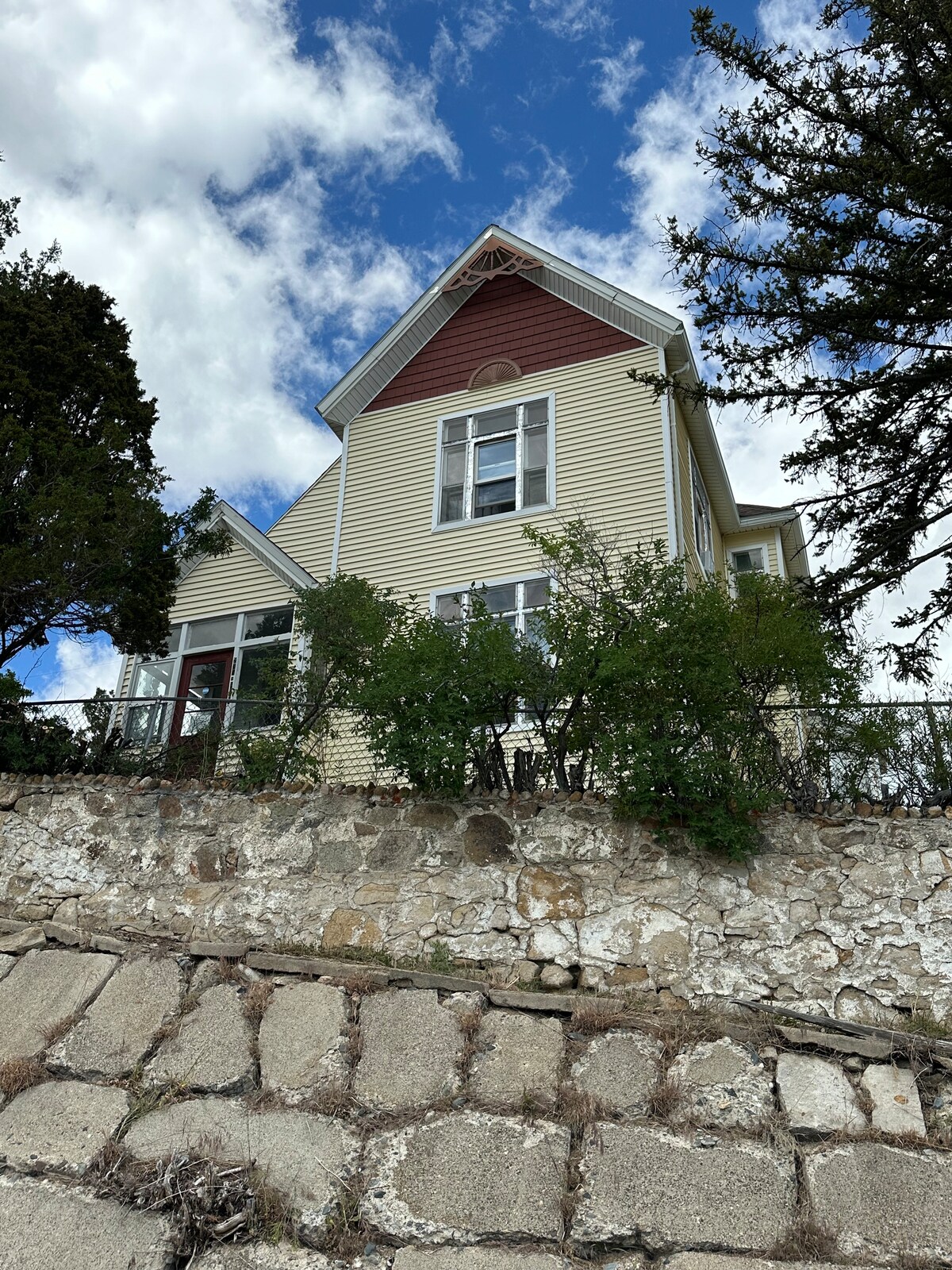 The large Victorian house is prominently situated on a hillside, showcasing a combination of yellow and red siding. Large windows allow for plenty of natural light, surrounded by greenery and the stone wall in the foreground adds an element of texture.