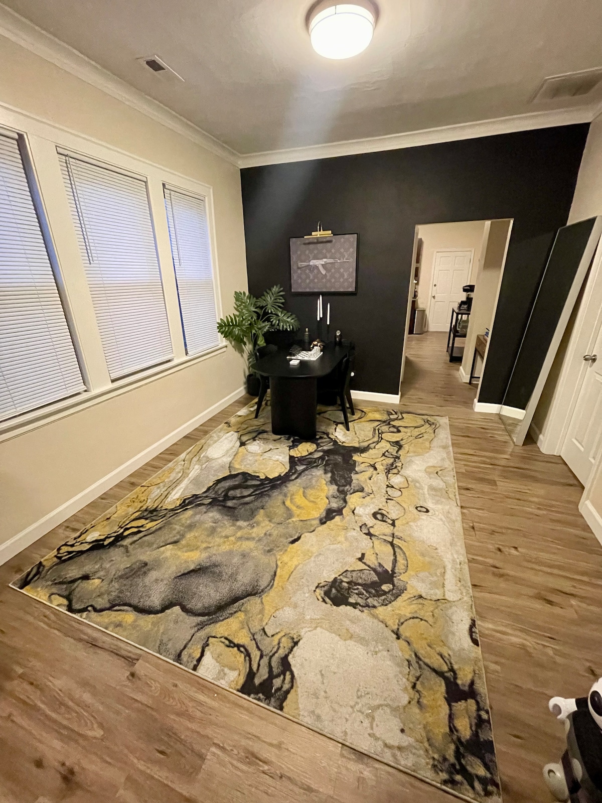 A corner of a room is highlighted, featuring a round black dining table surrounded by four chairs. An area rug with abstract patterns in yellow and gray covers the wooden floor. Large windows with blinds provide natural light, while a piece of wall art adds a touch of decor.