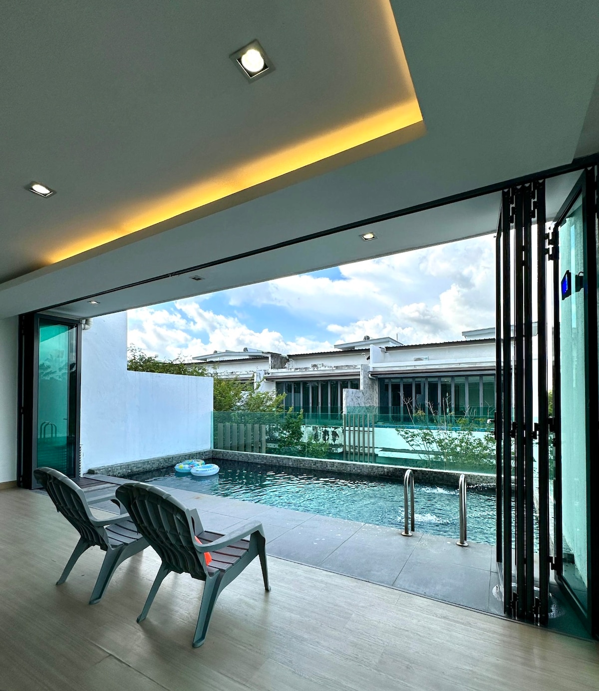Large glass doors open to a private pool area, with two charcoal gray lounge chairs positioned for relaxation. The pool reflects the building's modern architecture, surrounded by lush greenery and a clear sky overhead.