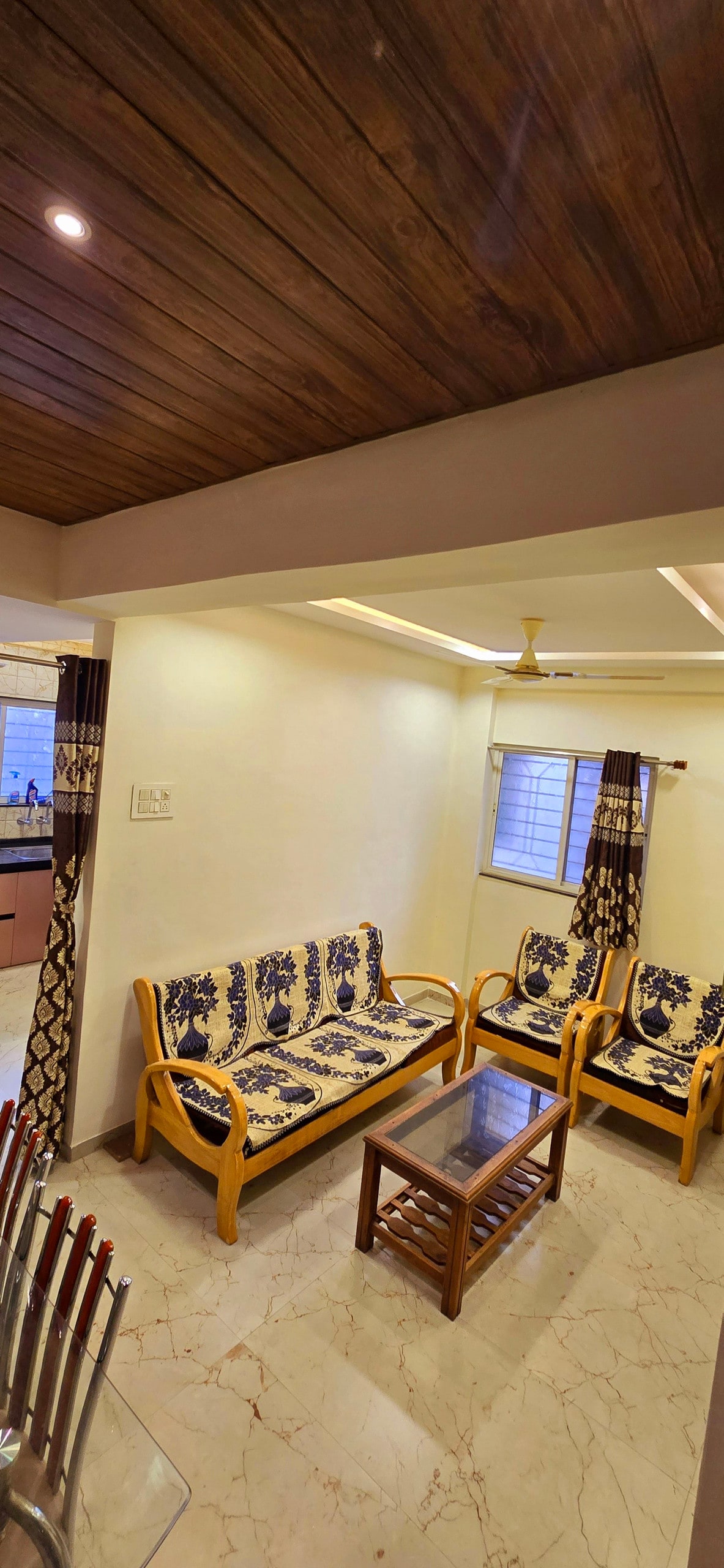 A comfortable seating area features a set of two floral-patterned sofas and a wooden coffee table. Natural light enters through a nearby window covered with patterned curtains, illuminating the polished marble flooring and the wooden ceiling above.