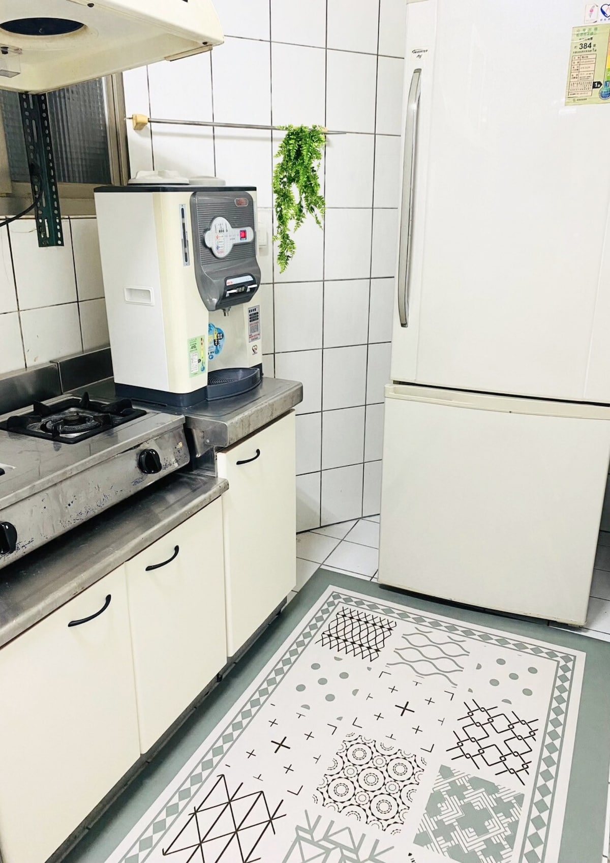 A compact kitchen is equipped with a gas stove and a refrigerator. A water dispenser is positioned next to the kitchen counter. The tiled walls and clean layout create an organized space, highlighted by a patterned rug on the floor.