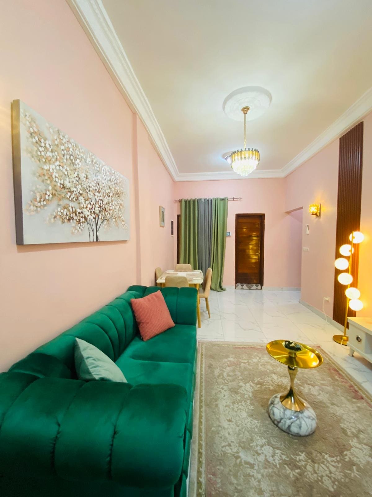 A living area features a green velvet sofa with accent cushions, complemented by a soft area rug. A round coffee table with a marble base sits in front. A dining space with wooden chairs is visible in the background, under soft lighting from an elegant chandelier.